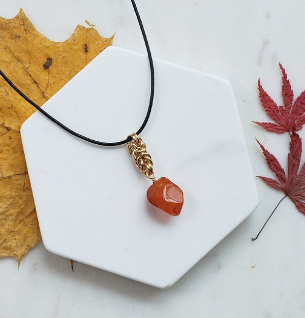 Glossy carnelian chunky and unfaceted is wire wrapped and hangs from a golden segment in byzantine pattern. The pendant hangs from a black leather cord.