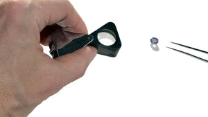 a hand holding a jewelers loupe a gemstone lying on white background with diamond tweezers