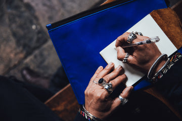image for kbeau blog post about why jewelry turns your skin green black. The image is a man's hands. He's wearing lots of big silver rings, some have gemstones. And he is writing in a small journal.