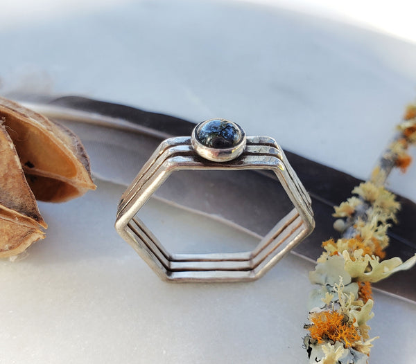 Three hexagonal rings soldered together to create one wide band. One hematite stone is centered on top. The background is lichen, a feather and seed pod, all on a light blue background.