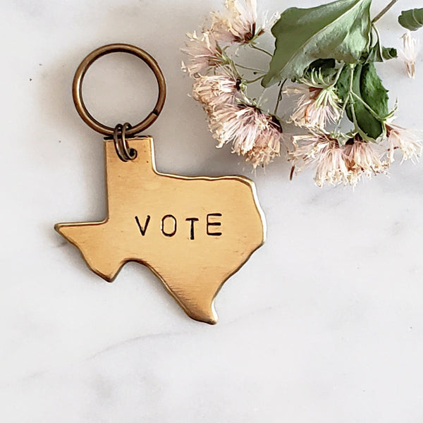 Texas-shaped keychain of brass with the word Vote hand stamped on it. The background is white marble and a dried white flower lies nearby