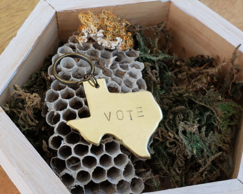 texas shaped brass keychain hand stamped with the word vote. background is moss, lichen and wasp nest