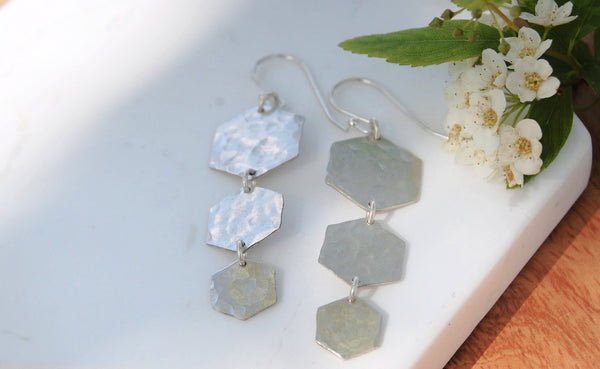 Sterling silver earrings on white marble background with small white flowers around it. Each earring has three tiered hexagons lightly hammered for texture.